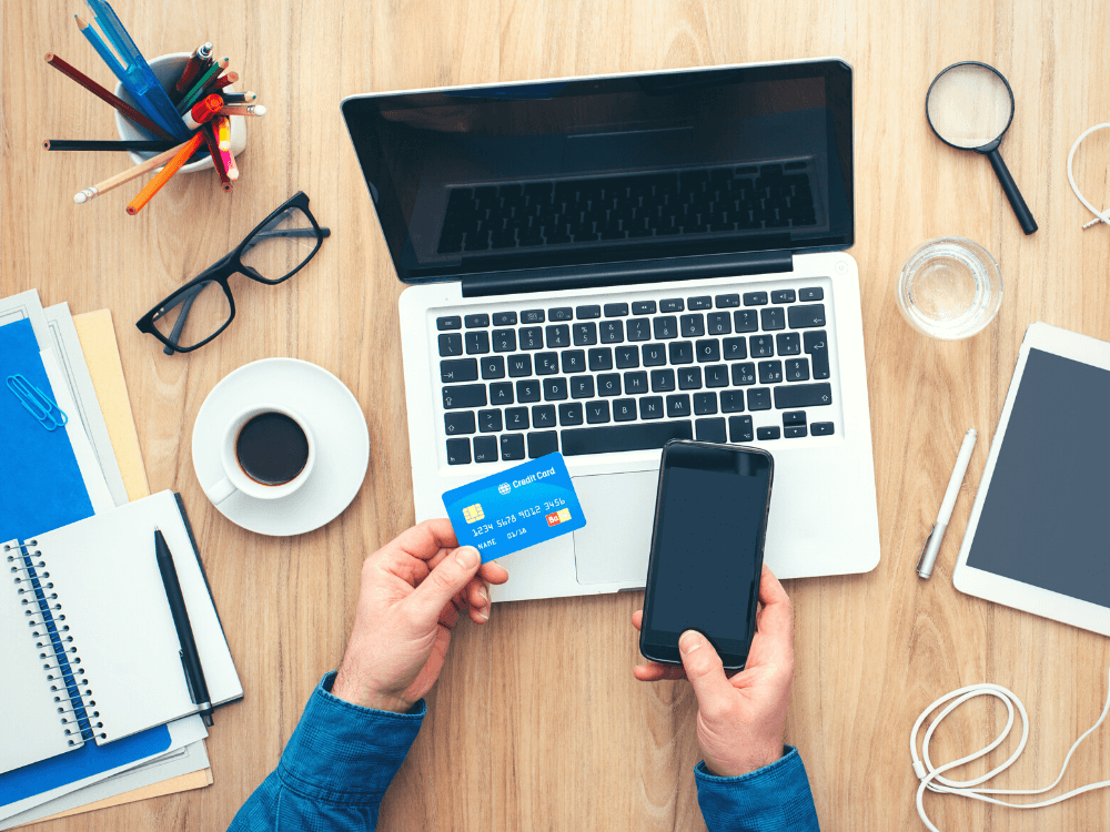 Pagamento no E-commerce: imagem de mãos vistas de cima segurando cartão de crédito e mexendo no celular. Mesa de madeira simples, laptpp e itens de escritório ao redor.