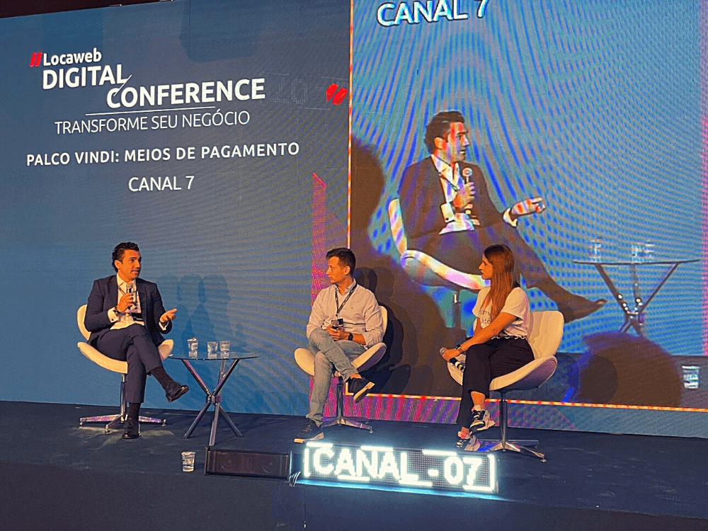 Locaweb Digital Conference: imagem da segunda palestra do palco vindi. Três pessoas sentadas em cadeiras na frente de um telão conversando.