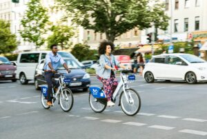 Mulher e homem pedalam em avenida movimentada de grande centro urbano