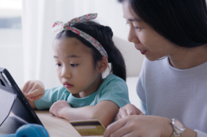 Mensalidades em Dia na educação:. Mãe e filha asiáticas olhando atentas algo em um tablet. A menina, que aparenta ter 4 ou 5 anos, usa laço rosa na cabeça;