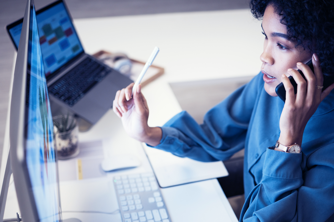 Código MCC - empreendedora negra sentada na frente do computador fala ao telefone enquanto confere planilha.