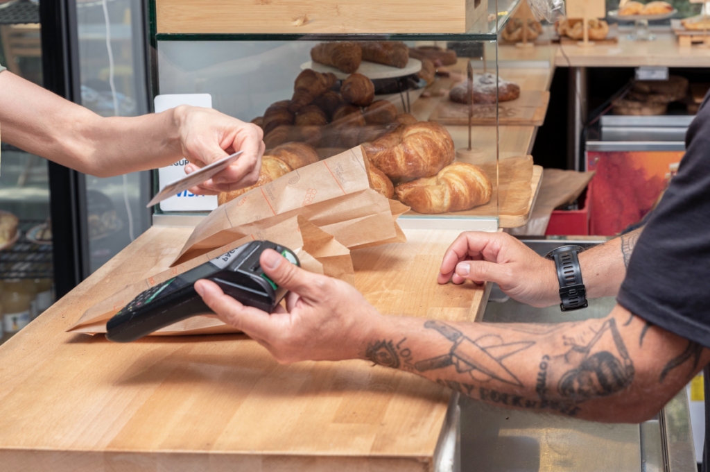 Código MCC - pessoa realizando pagamento em padaria via cartão de crédito. É possível observar parte do balcão e as mãos do vendedor e do pagador. 